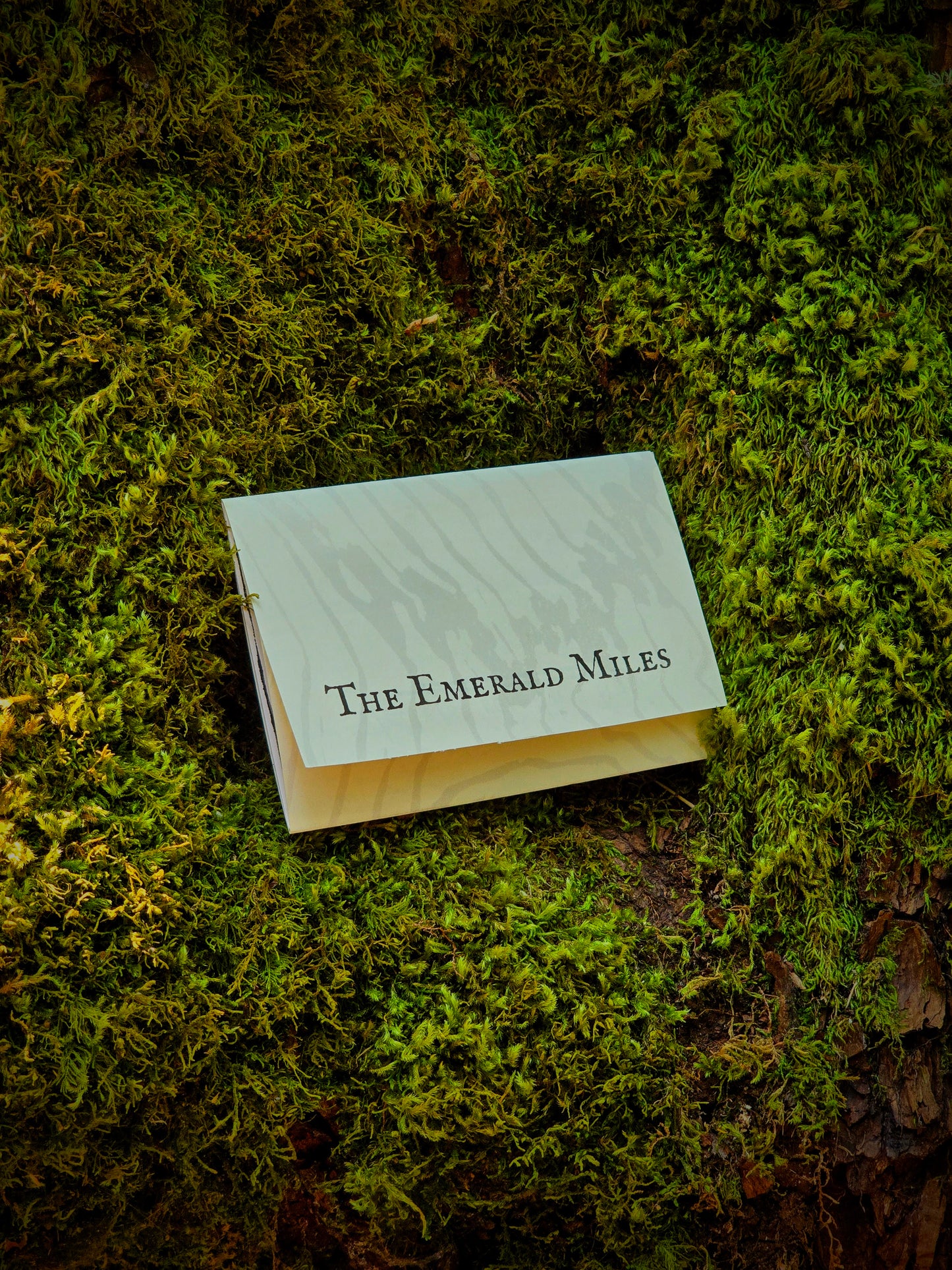 A small white rectangular card or booklet with the text 'The Emerald Miles' printed in black serif font, placed on a bed of green moss outdoors.