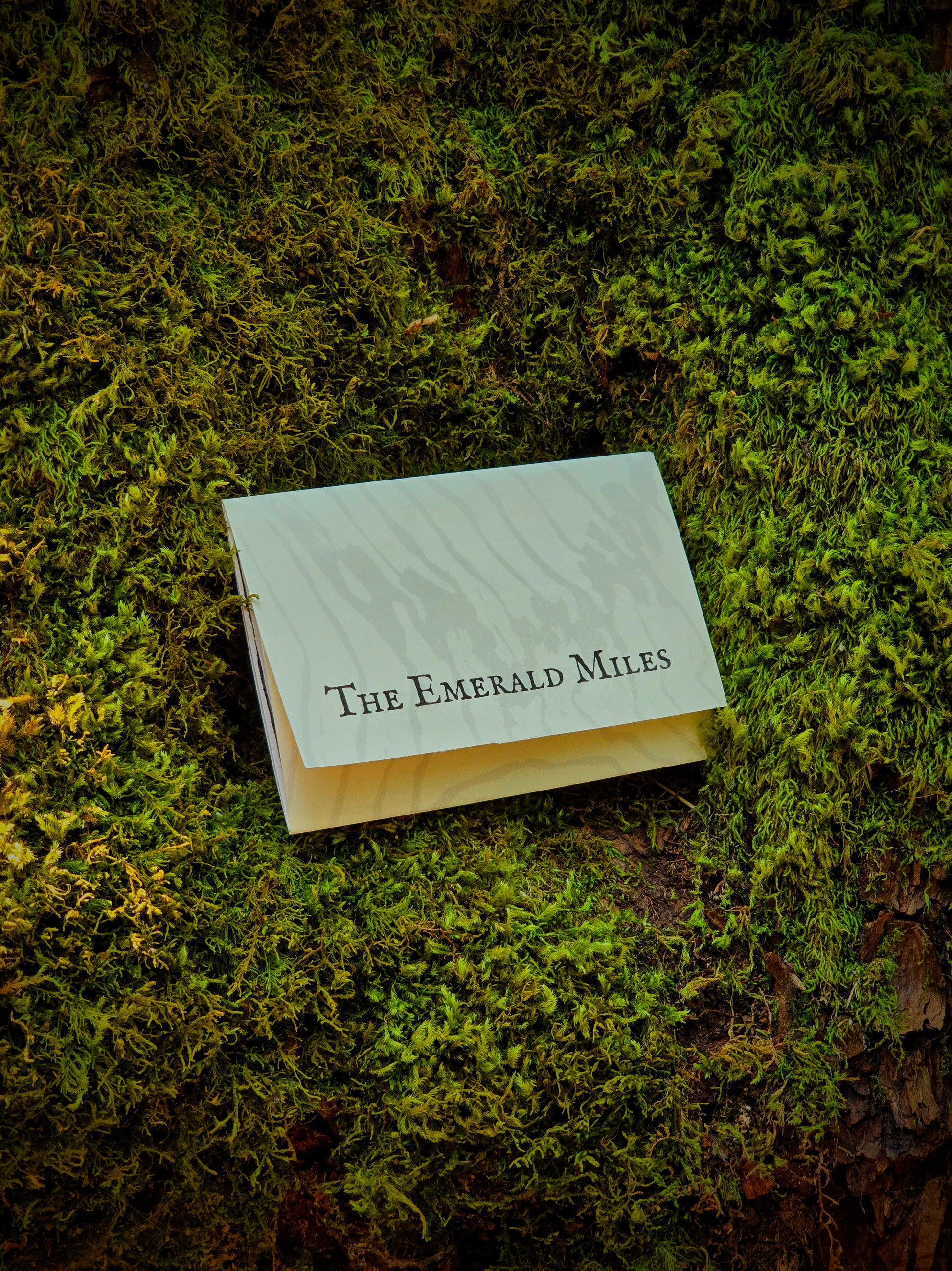 A small white rectangular card or booklet with the text 'The Emerald Miles' printed in black serif font, placed on a bed of green moss outdoors.