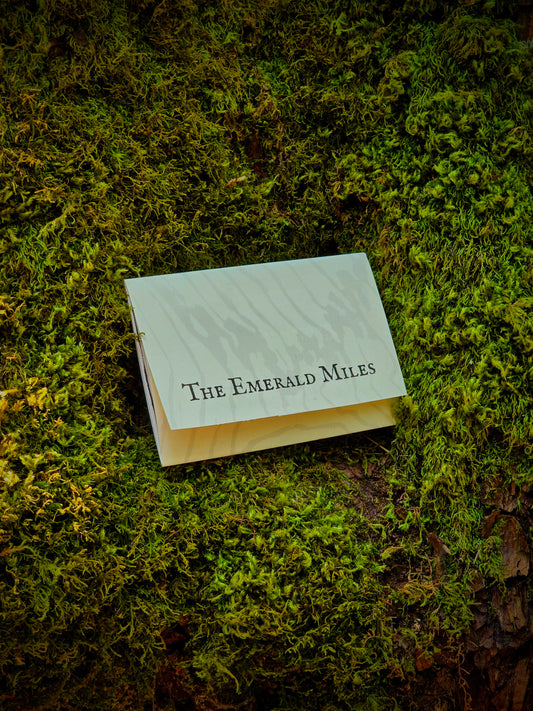 A small white rectangular card or booklet with the text 'The Emerald Miles' printed in black serif font, placed on a bed of green moss outdoors.
