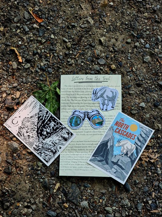 Three printed cards or postcards laid out on a gravel surface outdoors. The center card is titled "Letters from the Trail" and shows an elephant and binoculars illustration.
