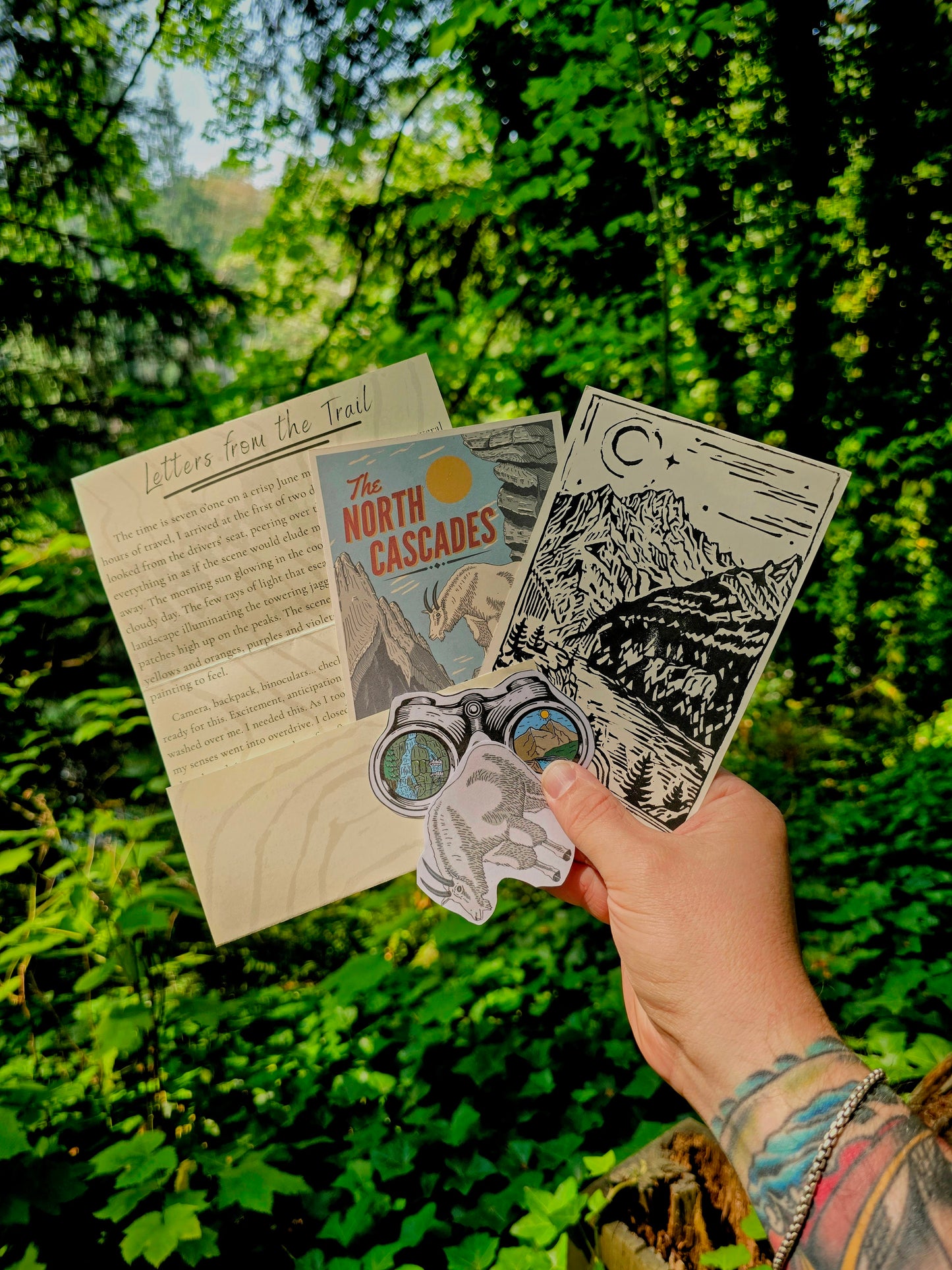 A hand holds a set of outdoor-themed stickers and printed cards in front of a lush green forest background. The set includes a letter from the trail, a card titled 'The North Cascades', a black-and-white mountain illustration card, and a die-cut sticker of a mountain goat wearing binoculars.
